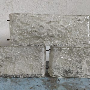 May include: Three rectangular, clear glass blocks with a textured, ice-like appearance. The blocks are arranged in a stacked configuration, with two blocks at the bottom and one on top. The glass has a slightly frosted look, and the edges are rough.
