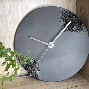 Può includere: Un orologio rotondo in cemento grigio con lancette bianche. Il quadrante dell'orologio ha una superficie strutturata ed è decorato con accenti maculati bianchi e neri. L'orologio è esposto su una mensola di legno accanto a una pianta in vaso.