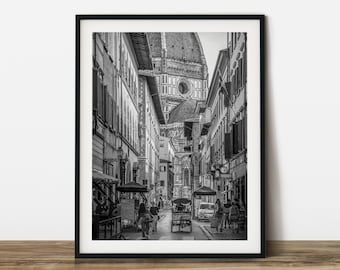 May include: Black and white framed print of a narrow street in Florence, Italy, with buildings lining both sides. The Duomo's dome is visible at the end of the street, and people are walking along the sidewalk. A small vehicle is in the middle of the street.