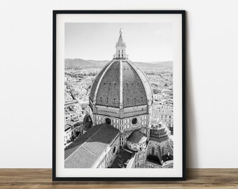 May include: Black and white framed print of the Florence Cathedral, Italy. The image captures the dome and surrounding cityscape from an aerial perspective. The print is in a black frame and is displayed on a wooden surface.