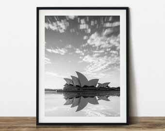 May include: Black and white framed print of the Sydney Opera House, a prominent landmark. The image captures the building's unique sail-like structure reflected in the water, under a cloudy sky. The print is displayed in a black frame.