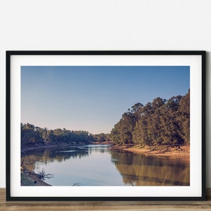 Op de afbeelding: Een ingelijste landschapsafdruk met een serene rivierscène. De afbeelding toont een kalme rivier die de omringende bomen en een heldere blauwe lucht weerspiegelt. Het kunstwerk wordt gepresenteerd in een zwarte lijst, wat een vleugje elegantie toevoegt.