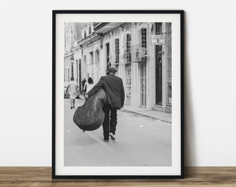 Può includere: Stampa incorniciata in bianco e nero di una scena di strada. Una persona con giacca e cappello cammina per una strada acciottolata portando un grande contrabbasso. Altre persone camminano sullo sfondo vicino agli edifici.
