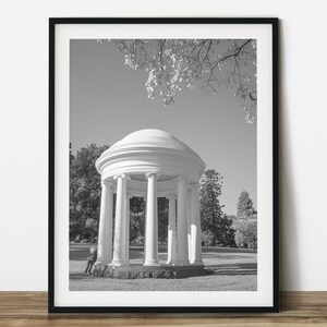 May include: Black and white framed print of a classical rotunda with columns, set in a park. The image captures the architectural details of the structure, with trees and sky in the background. A person is sitting on the base of the rotunda.