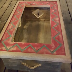 Op de afbeelding: Een houten doos met een rood en groen geometrisch patroon rond de bovenrand. De doos heeft een uitsparing in het midden en een kleinere uitsparing aan de onderkant.