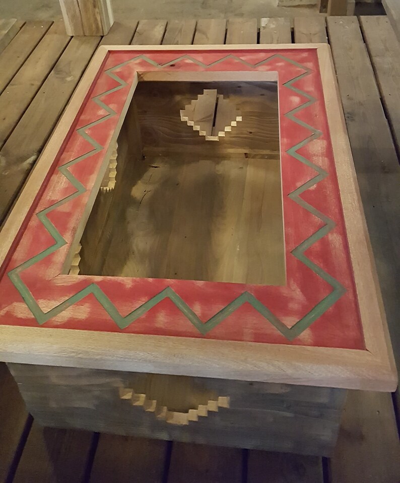 Op de afbeelding: Een houten doos met een rood en groen geometrisch patroon rond de bovenrand. De doos heeft een uitsparing in het midden en een kleinere uitsparing aan de onderkant.