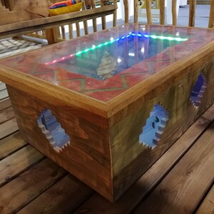 Op de afbeelding: Een houten salontafel met een glazen blad en een rood en oranje tegelontwerp rond de rand. Het glazen blad laat een blauw, groen en rood licht doorschijnen. De tafel heeft twee openingen aan de zijkanten, die een blauw licht in het interieur onthullen.
