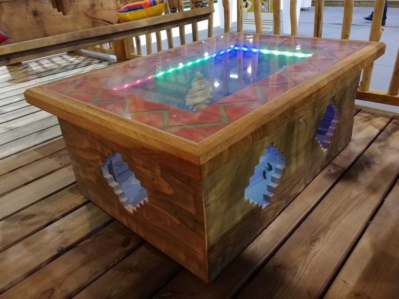 Op de afbeelding: Een houten salontafel met een glazen blad en een rood en oranje tegelontwerp rond de rand. Het glazen blad laat een blauw, groen en rood licht doorschijnen. De tafel heeft twee openingen aan de zijkanten, die een blauw licht in het interieur onthullen.