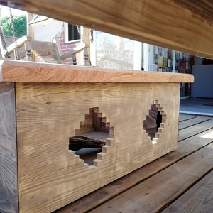 Op de afbeelding: Een houten doos met twee uitgesneden vormen. De doos is gemaakt van lichtbruin hout en heeft een natuurlijke afwerking. De uitgesneden vormen zijn in de vorm van een ruit en een cirkel. De doos staat op een houten dek.