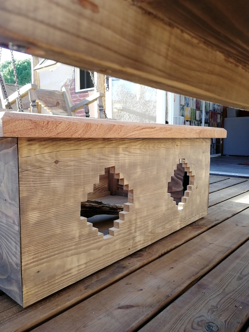 Op de afbeelding: Een houten doos met twee uitgesneden vormen. De doos is gemaakt van lichtbruin hout en heeft een natuurlijke afwerking. De uitgesneden vormen zijn in de vorm van een ruit en een cirkel. De doos staat op een houten dek.
