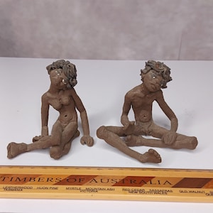 May include: Two brown clay figurines of seated figures with textured hair and defined facial features. The figures are positioned behind a wooden ruler labelled Timbers of Australia, which shows a scale in inches and centimetres for size reference.