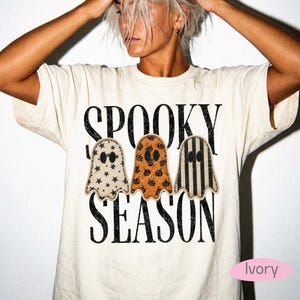 May include: An ivory short-sleeve t-shirt featuring the black distressed text SPOOKY SEASON. Below the text are three ghost-shaped graphics: one with black stars, one with orange pumpkins, and one with black and ivory vertical stripes.