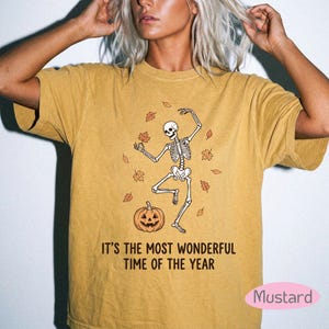 May include: A mustard yellow short-sleeve t-shirt featuring a graphic of a dancing skeleton surrounded by falling autumn leaves and a jack-o'-lantern. The text "IT'S THE MOST WONDERFUL TIME OF THE YEAR" is printed in black below the graphic.