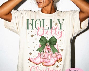 May include: An ivory t-shirt featuring a graphic print with the text Holly Dolly Christmas in green and pink. The design includes two pink cowboy boots adorned with a large green bow and small gold stars scattered across the front of the shirt.