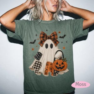 May include: A moss green short-sleeve t-shirt featuring a central graphic of a ghost wearing a plaid bow. The ghost has patchwork-style patterns on its base and carries an orange jack-o'-lantern pumpkin. Small black bats and stars surround the ghost.