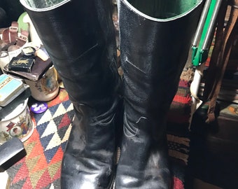 May include: A pair of tall, black leather boots with a shiny finish. The boots have a green interior and are set on a patterned rug. The boots appear to be vintage or antique, with visible wear and tear.