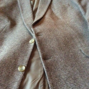 Puede incluir: Una blazer de tweed marrón con solapa de muesca y cierre de dos botones. La blazer tiene un forro marrón claro y dos bolsillos delanteros. Los botones son de color marrón claro. La blazer está hecha de una tela texturizada.