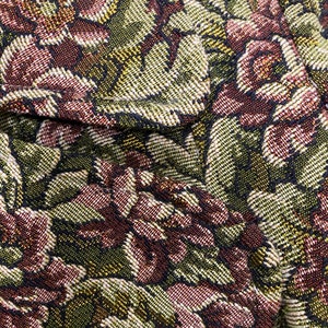 Op de afbeelding: Close-up van een geweven stof met een bloemenpatroon. Het ontwerp toont een herhalend patroon van gestileerde bloemen in tinten bordeaux, groen en goud, tegen een donkere achtergrond. De stof heeft een getextureerde uitstraling.