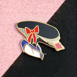 May include: Gold-tone enamel pin featuring a cartoon character wearing a black and white sailor hat with a red bow, a blue and white sailor collar, and a white shirt. The character is holding a red playing card with white stripes.