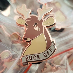 May include: Rose gold enamel pin featuring a cartoon deer with antlers and a banner that reads "Buck Off".