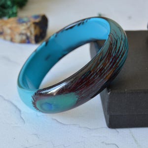 Puede incluir: Una pulsera rígida lisa y redondeada con un interior azul turquesa vibrante y una superficie exterior brillante decorada con un patrón de ojo de pluma de pavo real en marrón oscuro y verde que envuelve la circunferencia de la banda circular.