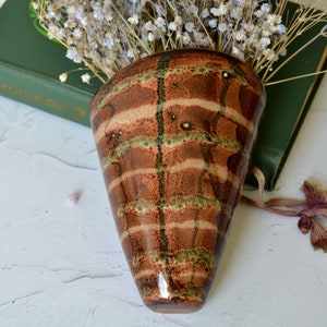 May include: A cone-shaped ceramic wall vase featuring a glossy brown glaze with horizontal bands of mottled green and tan. The surface has a smooth, reflective finish with a distinct grid-like pattern created by dark vertical and horizontal lines.