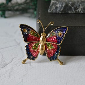 Puede incluir: Un broche de mariposa dorado con alas coloridas y brillantes. Las alas presentan un degradado de rojo, verde y azul, con un borde azul oscuro. El broche tiene un cuerpo y antenas detallados, y es un accesorio decorativo.