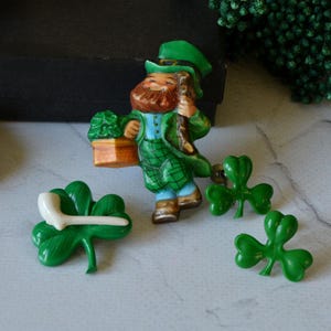 Op de afbeelding: Een verzameling St. Patrick's Day-thema pins en broches. De set bevat een leprechaun-beeldje met een pot goud, een klaverblad-pin met een witte pijp en drie groene klaverblad-pins. De leprechaun draagt een groene hoed en jas.
