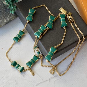 May include: A gold-toned necklace featuring a series of green bow-shaped charms. The bows have a glossy finish and are outlined in gold. The necklace is displayed on a dark surface, showcasing its delicate chain.