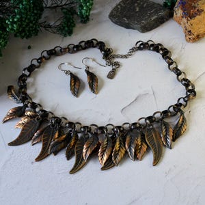 May include: A necklace and earrings set featuring a chain with dangling leaves. The leaves are made of metal and have a copper and black finish.