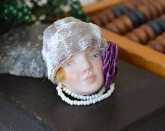 Puede incluir: Un broche con forma de cabeza de muñeca de porcelana con rostro pálido y rasgos pintados, que lleva un sombrero cloche gris claro texturizado, una flor de tela morada en el lateral y un collar de doble hilera de pequeñas cuentas blancas.