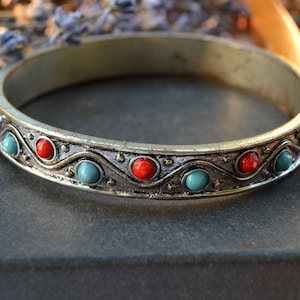 Può includere: Un braccialetto rigido argentato con una fascia decorativa con pietre rosse e turchesi alternate. Il braccialetto ha un aspetto vintage, con una finitura leggermente ossidata.