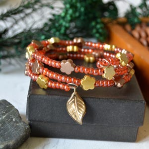 Könnte beinhalten: Ein mehrreihiges Wickelarmband aus durchscheinenden orangefarbenen Saatperlen, die mit kleinen, matten goldfarbenen blütenförmigen Perlen durchsetzt sind. Ein einzelner metallischer blattförmiger Anhänger hängt an der Mitte des kreisförmig gewickelten Armbands.