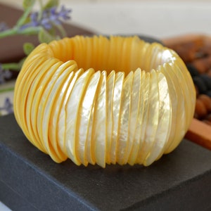 Puede incluir: Un brazalete de concha amarillo hecho de muchas conchas individuales. El brazalete está sobre una superficie negra.