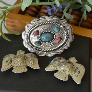 Puede incluir: Un conjunto de tres piezas de joyería plateadas con piedras turquesa y coral. La pieza central es un óvalo con un borde de cuerda y diseños geométricos. Las otras dos piezas son pájaros estilizados.