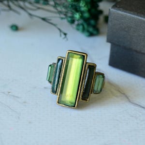 Puede incluir: Un anillo con una piedra central rectangular vertical de color verde claro, flanqueada por dos piedras rectangulares de color verde oscuro a cada lado, montadas en un marco metálico con acabado en tono bronce y diseño geométrico.