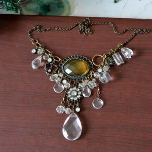 Peut inclure: Un collier imposant avec une pierre centrale ovale de couleur ambre montée sur une monture en métal bronze antique, orné de multiples gouttes de cristal transparent à facettes, de perles rondes et d'une breloque florale sur une chaîne assortie.