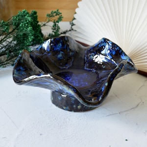 Op de afbeelding: Een kleine, met de hand gevormde keramische kom met een golvende, geplooide rand. Het object heeft een glanzend, diepblauw glazuur met gevlekte zwarte en donkerblauwe accenten over het gehele oppervlak en rust op een stevige, ronde voet.