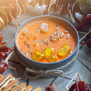 May include: Orange scented candle in a gray tin with a lid. The candle is decorated with dried orange peels, cinnamon sticks, red chili peppers, and dried flowers. The candle is surrounded by a brown twine bow.
