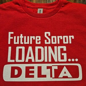K&ouml;nnte beinhalten: Rotes T-Shirt mit wei&szlig;em Glitzertext, der "Future Soror Loading... DELTA" lautet.