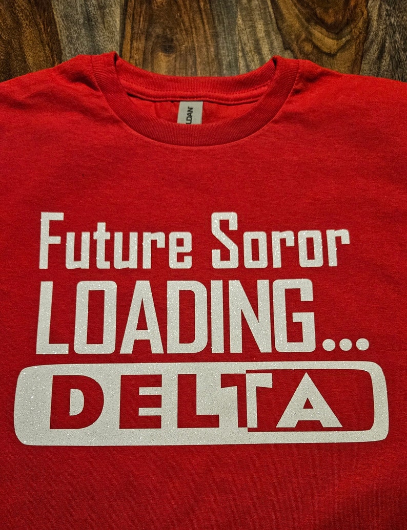 K&ouml;nnte beinhalten: Rotes T-Shirt mit wei&szlig;em Glitzertext, der "Future Soror Loading... DELTA" lautet.