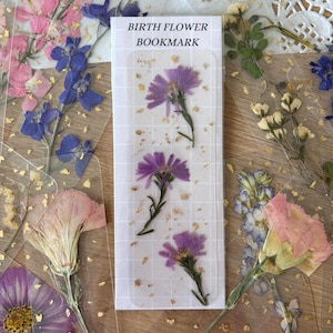 May include: A clear bookmark with three pressed purple flowers and gold flecks. The bookmark has the text "BIRTH FLOWER BOOKMARK" in black. Surrounding the bookmark are other pressed flowers in pink, purple, and blue.