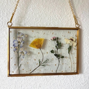 May include: A rectangular, gold-framed pressed flower art piece. The clear glass displays dried flowers in shades of yellow, purple, and white, with gold leaf accents. A gold chain is attached for hanging.