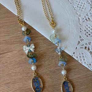 May include: Two hanging ornaments featuring gold-tone chains, white faux pearls, green and blue flower-shaped beads, and a white bow charm. Each ornament terminates in an oval gold-tone pendant containing pressed blue forget-me-not flowers.