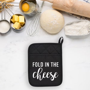 May include: Black oven mitt with white text that reads "Fold in the cheese". The oven mitt is on a white marble surface with various baking ingredients, including flour, sugar, eggs, butter, and dough.