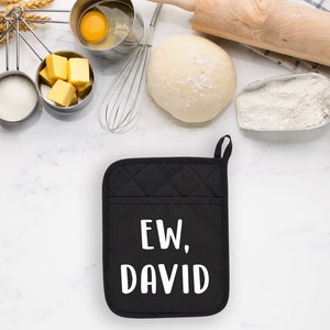 May include: Black quilted oven mitt with the text "EW, DAVID" in white. Baking ingredients and tools surround the mitt, including eggs, flour, butter, and a rolling pin. The scene is set on a white marble surface.