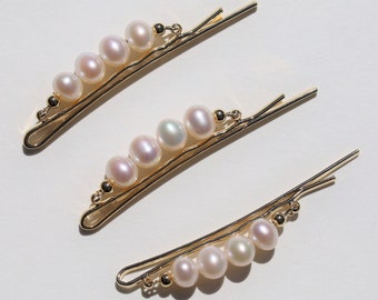 Puede incluir: Tres pinzas para el pelo doradas con detalles de perlas. Cada pinza tiene cinco perlas dispuestas en una línea curva.