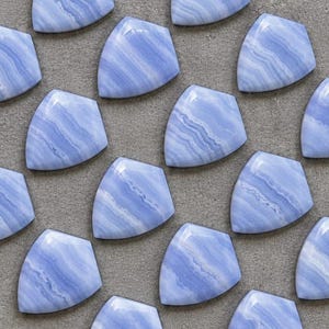 Könnte beinhalten: Eine Sammlung von schildförmigen Cabochons aus blauem Spitzenachat, die jeweils ausgeprägte hellblaue und weiße wellenförmige Bänder aufweisen. Diese polierten Steine sind in einem Raster angeordnet und zeigen ihre glatten Oberflächen.