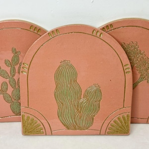 May include: Three ceramic coasters with a coral pink background and a gold trim. Each coaster features a different desert plant design in green and gold. The designs include a cactus, a tree, and a prickly pear.