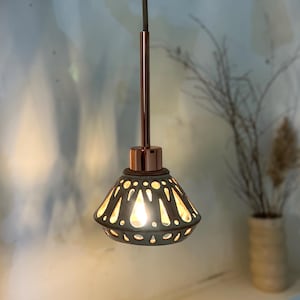 May include: A pendant light fixture with a copper-colored stem and a dark gray, cone-shaped shade. The shade has a pattern of cut-out shapes that allow light to shine through. The light is hanging from a dark cord.